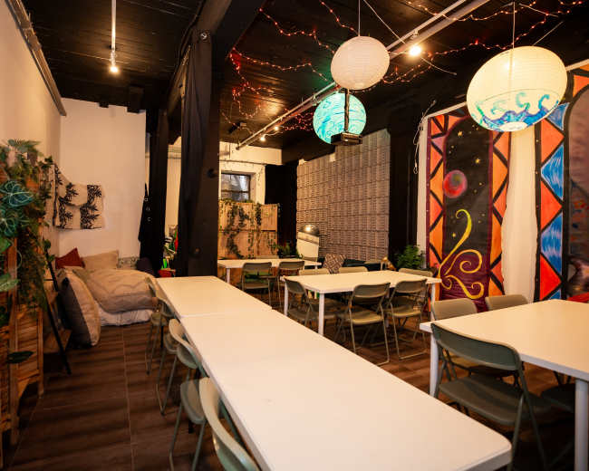 The image shows a brightly lit communal space featuring several long tables, chairs, and decorative wall art, with plants scattered throughout the room.