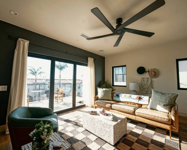 The image shows a modern living room with a large sliding glass door that leads to an outdoor patio, featuring a sofa, a coffee table, and a ceiling fan.