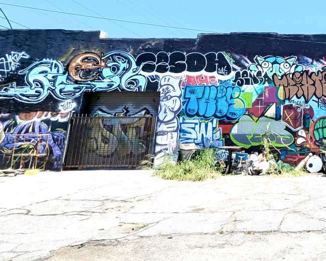 The image shows a large brick wall covered in vibrant graffiti art, with an assortment of discarded furniture and items in the foreground.