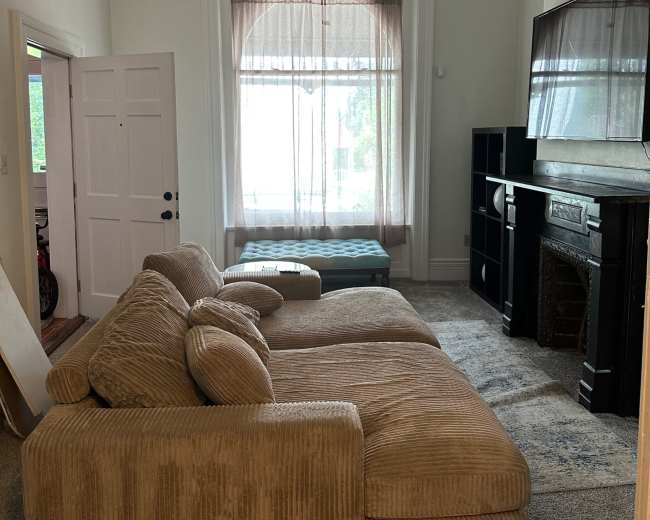 The image shows a cozy living room featuring a large sectional sofa, a TV mounted on the wall, and a window with sheer curtains letting in light.
