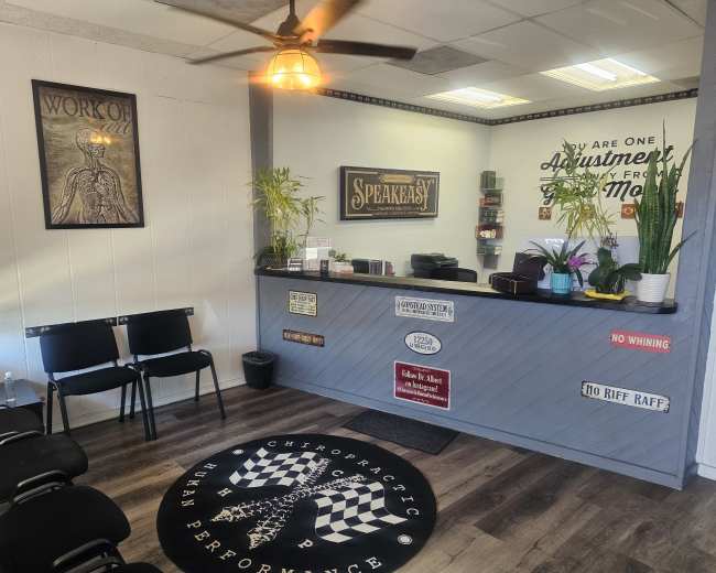 The image shows a chiropractic clinic reception area with a front desk, seating for visitors, and various wall decorations.