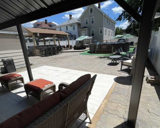 The image shows a patio area with a seating arrangement under a roof, overlooking a spacious outdoor space featuring a grassy area, parked vehicles, and shaded tables.