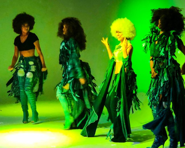 Four dancers with large hairstyles perform on stage while wearing vibrant, textured outfits under green lighting.