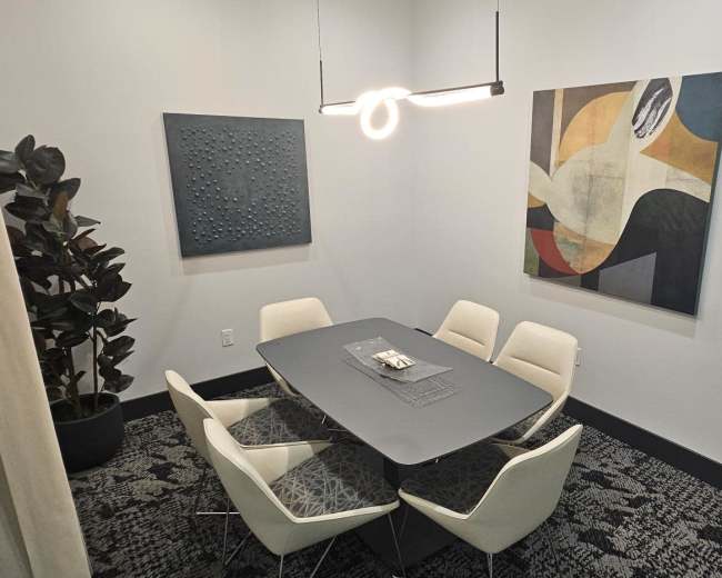 A modern conference room features a round table surrounded by white chairs, with a plant in the corner and abstract art on the walls.