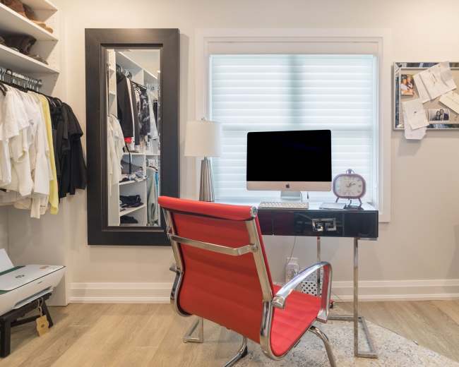 A modern home office is set up in a walk-in closet, featuring a desk with a computer, a red chair, and organized clothing on racks.