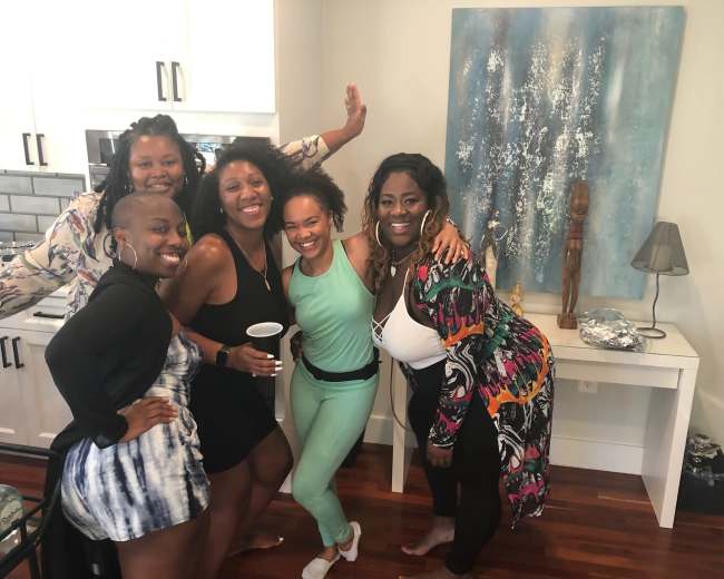 Five women gather in a kitchen, smiling and posing together in front of a light blue painting on the wall.