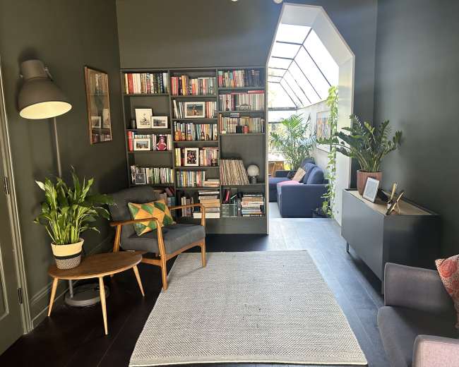 The image shows a cozy living space featuring a bookshelf, indoor plants, and comfortable seating arranged around a light-filled area with a glass roof.