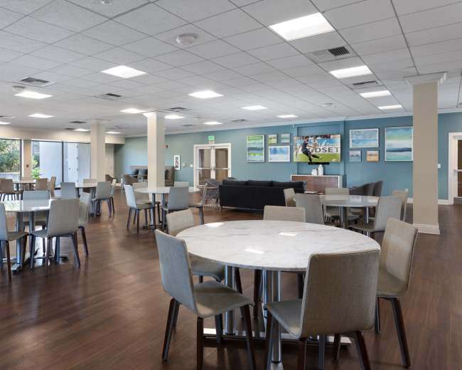 The image shows a spacious dining area with multiple round tables and chairs, featuring large windows that display a view of the outdoors.