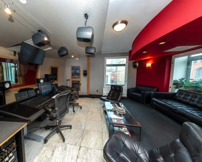 The image shows a music recording studio featuring a control desk, sound equipment, and seating areas with leather chairs and a sofa.