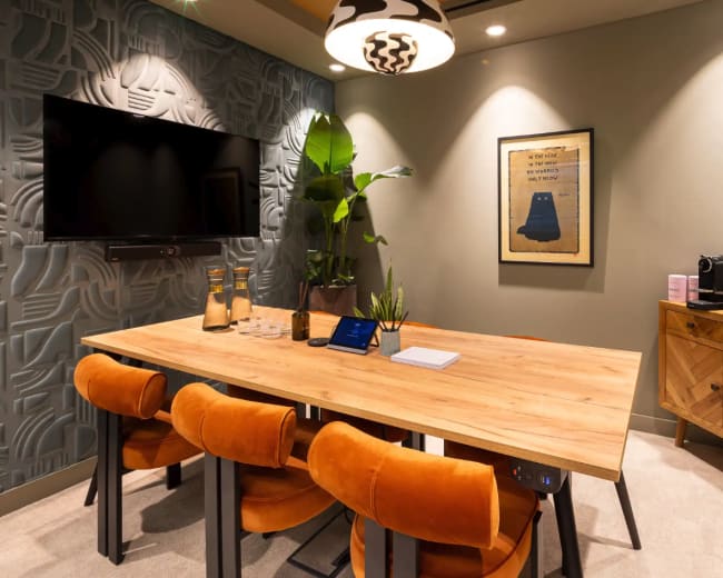 The image shows a small meeting room featuring a wooden table surrounded by orange chairs, a television mounted on the wall, and decorative items including a plant and artwork.