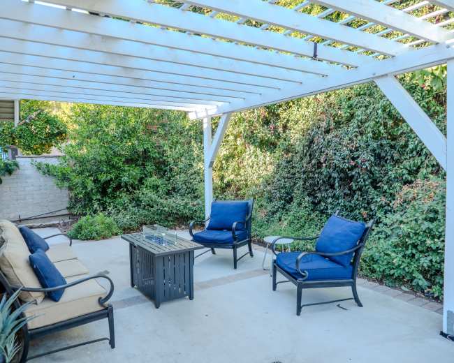 The image shows a patio area with a white pergola, outdoor seating, and a central table surrounded by greenery.