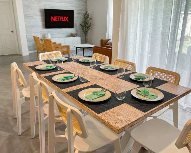 A dining table is set for eight with plates and glasses, and a television displaying the Netflix logo is mounted on the wall in the background.