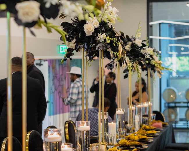 A long banquet table is set with black and gold tableware, elegant floral centerpieces, and candles, ready for an event.