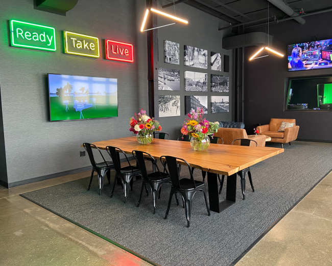 The image shows a modern conference room featuring a large wooden table surrounded by black chairs, with vibrant flower arrangements and neon signs on the wall, alongside large screens displaying images and a television setup.