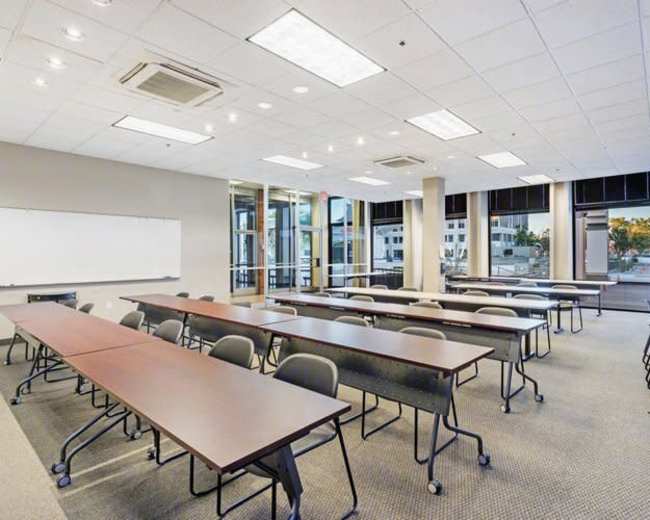 A bright classroom is set up with rows of long tables and chairs, featuring large windows that allow natural light to filter in.