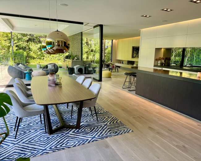 A modern dining area with a large table, stylish chairs, and an open kitchen overlooking a garden through large glass windows.