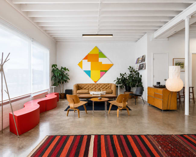 A modern living room features a brown sofa, a colorful geometric wall art piece, and a mix of seating surrounded by plants and wooden furniture.