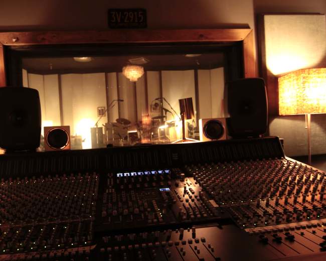 A recording studio control room is shown with a large mixing console in the foreground and a glass partition overlooking a soundproof room with instruments and lights.