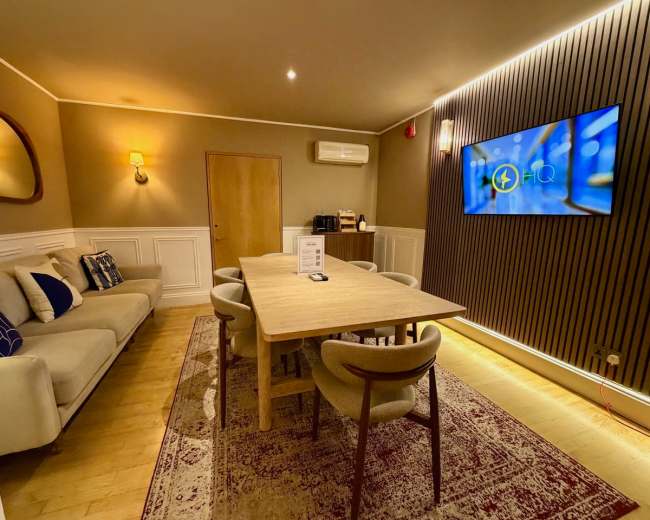 The image shows a well-furnished meeting room with a dining table surrounded by chairs, a sofa, and a large screen mounted on the wall.
