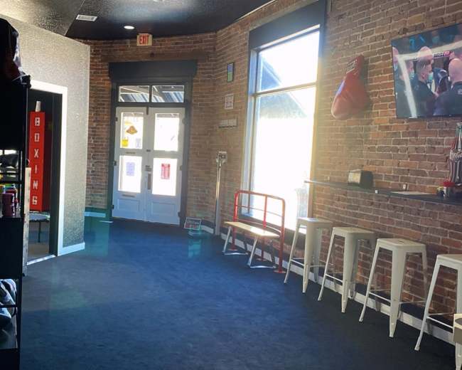 The image depicts a well-lit corridor of a gym with exposed brick walls, a boxing bag hanging, and seating along one side.