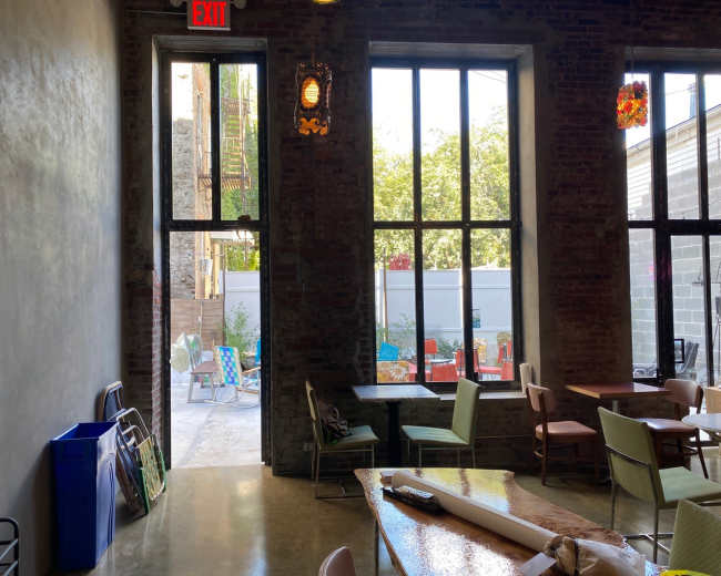 The image shows a spacious interior of a cafe or restaurant with large windows that open to an outdoor patio area, featuring tables and colorful chairs.