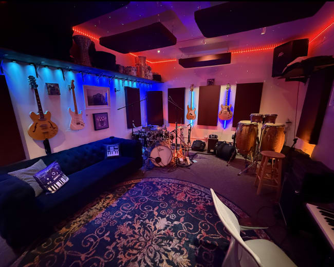The image shows a vibrant music studio with bright lighting, featuring guitars mounted on the walls, a drum set, conga drums, and a comfortable couch on a patterned rug.