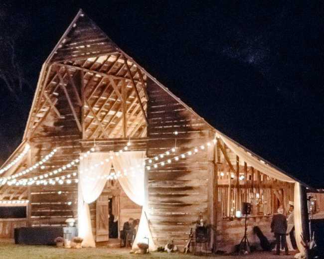 A barn is illuminated at night with string lights and curtains, creating a festive atmosphere.
