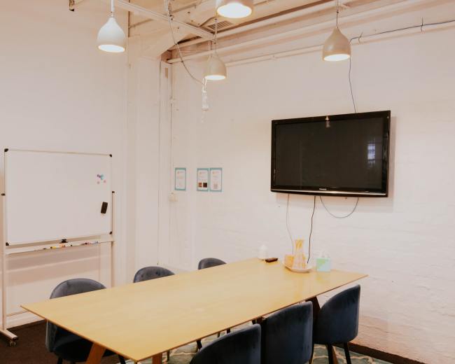 A meeting room features a long table surrounded by chairs, a whiteboard on wheels, and a mounted television on the wall.