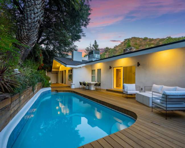 A modern house with a swimming pool and wooden deck is set against a hillside backdrop under a colorful sky.