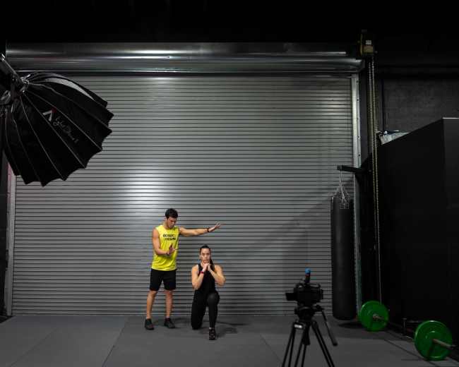 A man in a yellow tank top stands beside a woman in a squat position in a gym setting with fitness equipment visible.
