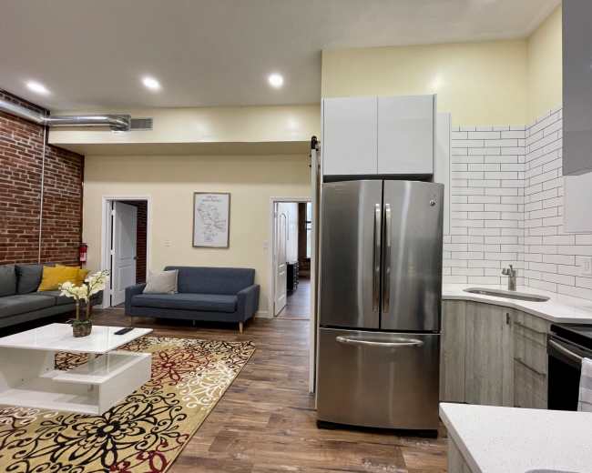 A modern kitchen and living area showcase a stainless steel refrigerator, a brick wall, and contemporary furniture.