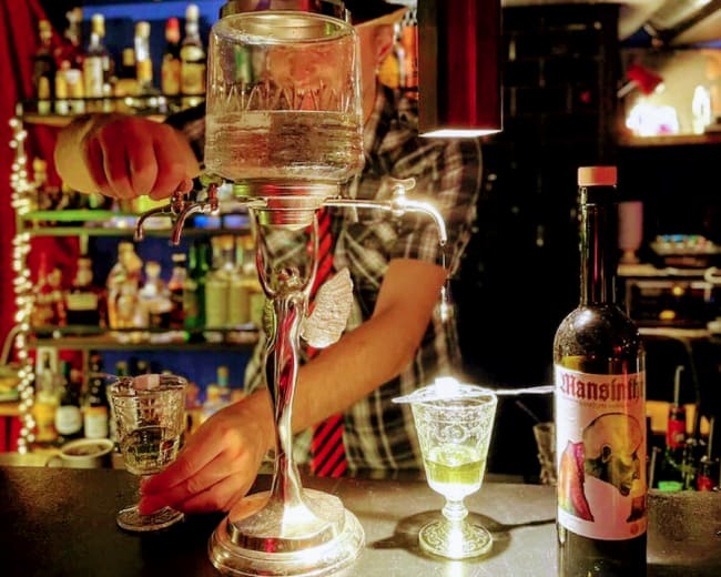 A bartender pours a clear liquid from a vintage dispenser into a glass at a dimly lit bar, with shelves of bottles in the background.