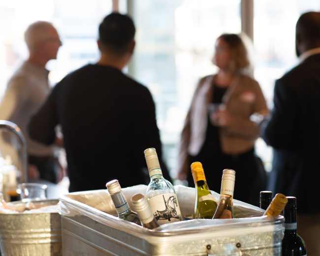 A metal bucket filled with various bottles of wine sits in the foreground while people chat in the background.