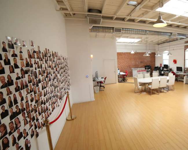 The image shows a large modern office space with a polished wooden floor, featuring a long table surrounded by white chairs, a wall covered with numerous photographs, and exposed brick walls.