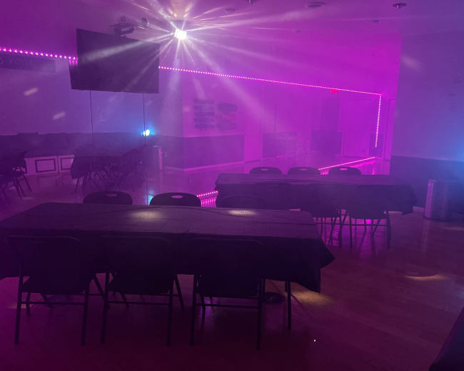 The image shows a dimly lit room with several black tables and purple lighting along the walls.