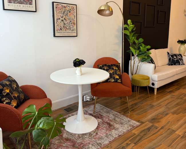 The image shows a stylish living room with a white sofa, orange chairs, a round table, decorative plants, and artwork on the walls.