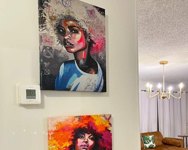 Two colorful paintings of women with distinctive hairstyles hang on a white wall in a living room featuring a modern chandelier and a round coffee table.
