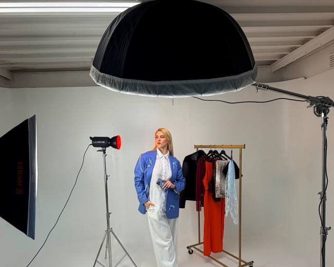 A model poses in a studio setting, standing in front of a clothing rack with a variety of garments while under bright studio lights.