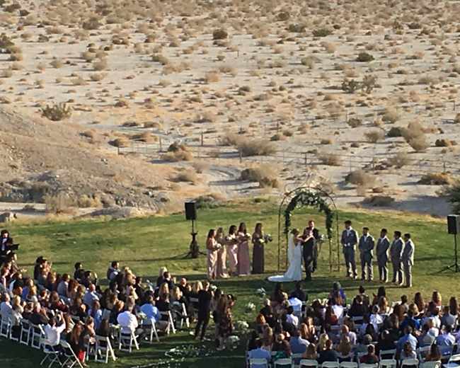 A couple stands under an arch during their wedding ceremony in a desert setting surrounded by guests seated in rows.