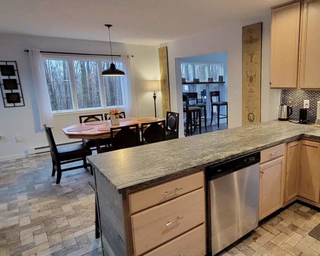 The image shows a modern kitchen with light wood cabinetry, a stainless steel dishwasher, and a view of an adjoining dining area with a round table and chairs.