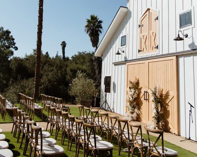 A wedding venue features a white barn with wooden doors, surrounded by palm trees, and lined with empty chairs for a ceremony.