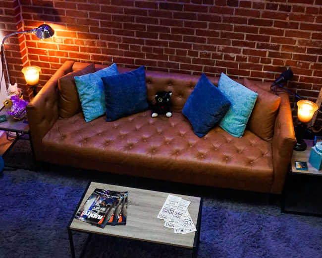 The scene features a brown leather couch with blue cushions against a brick wall, accompanied by a small coffee table and a few decorative items.