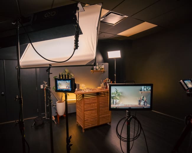 The image shows a studio setup with lights and cameras focused on a wood-paneled bar area equipped with various bottles and plants.