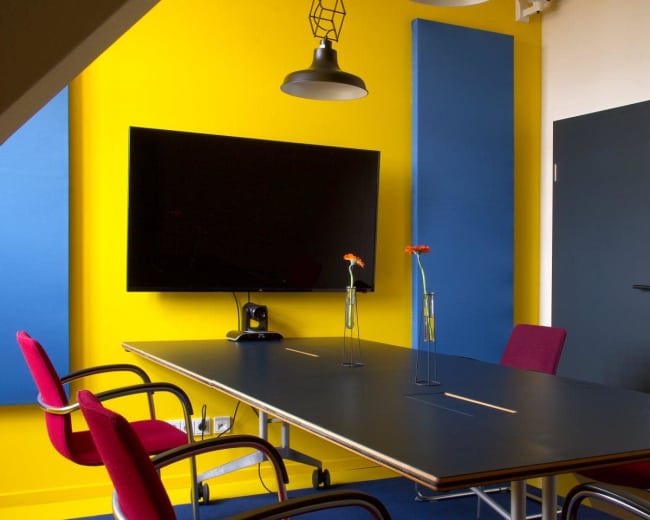 A modern conference room features a rectangular black table, colorful wall panels, a large television screen, and two pink chairs arranged around the table with small vases containing flowers.