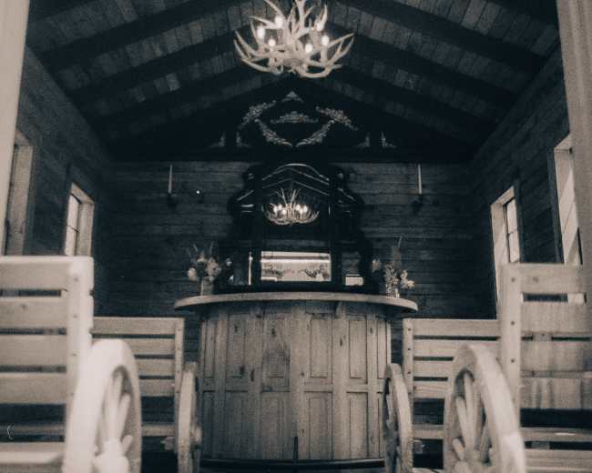 The image displays a rustic interior with wooden walls and ceiling, featuring a circular bar counter and benches arranged in a symmetrical layout.