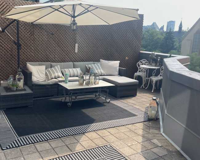 The image shows a rooftop terrace with a sectional sofa, a large umbrella, and a coffee table surrounded by decorative elements and cityscape in the background.