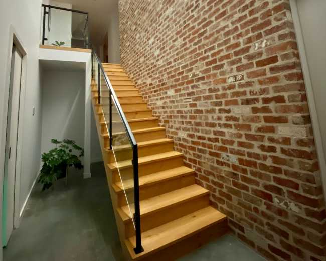 A wooden stairway leading to an upper level, flanked by a brick wall and illuminated by pendant lights and recessed lighting.