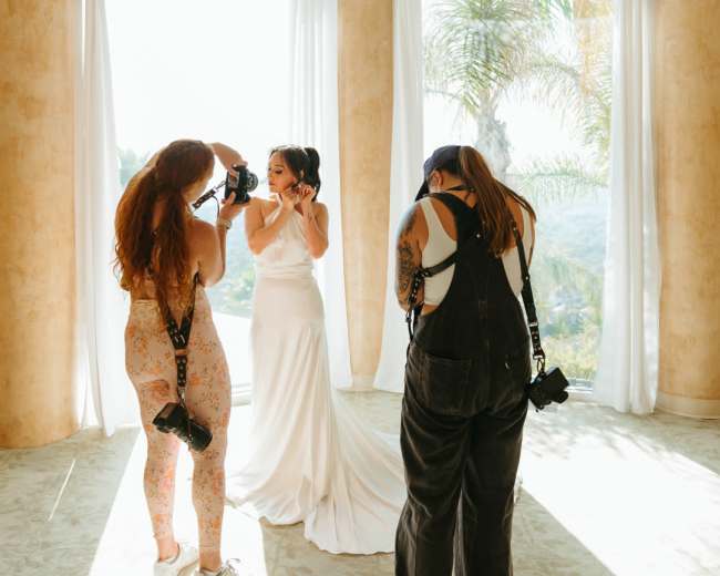 A bride is getting her makeup done while two women photographers capture the moment in a brightly lit room with large windows.