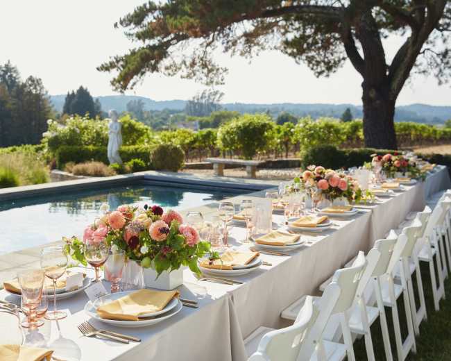 A long dining table is set outdoors beside a swimming pool, adorned with floral centerpieces, with rolling hills and vineyards in the background.