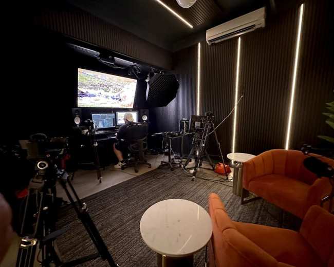 A person is seated at a desk with monitors in a modern studio equipped with cameras and orange chairs.